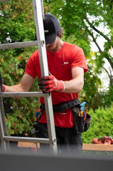 A gardener wearing safety gear climbs a ladder outdoors, surrounded by green foliage.